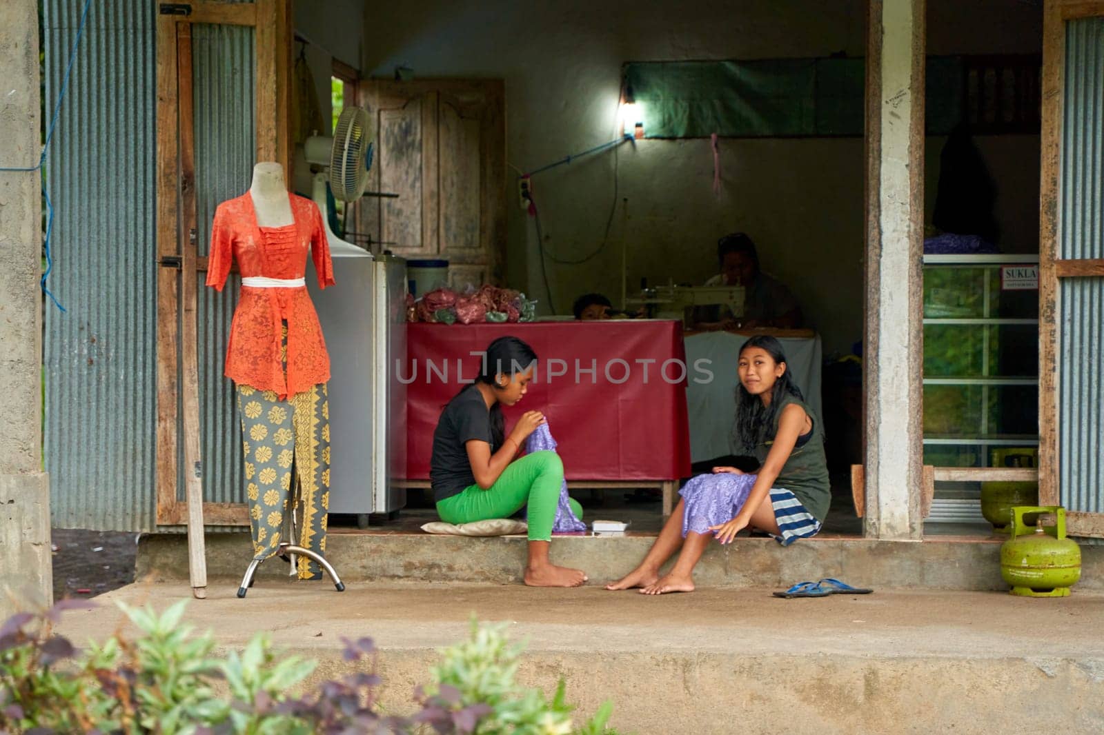 People resting near the road in an unusual Asian village. Children are playing and adults are talking sitting on the roadside. Bali, Indonesia - 12.24.2022