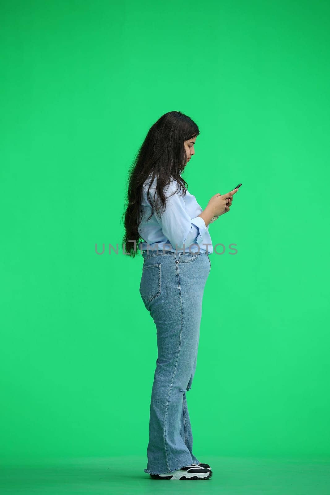 A girl in a blue shirt, on a green background, full-length, with a phone in her hands, in profile.