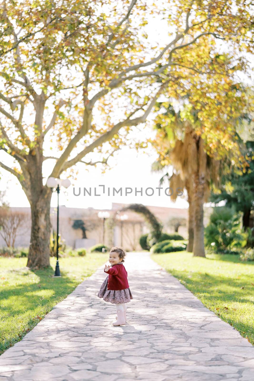 Little girl stands half-turned on a paved path in the park and waves her hand affably by Nadtochiy