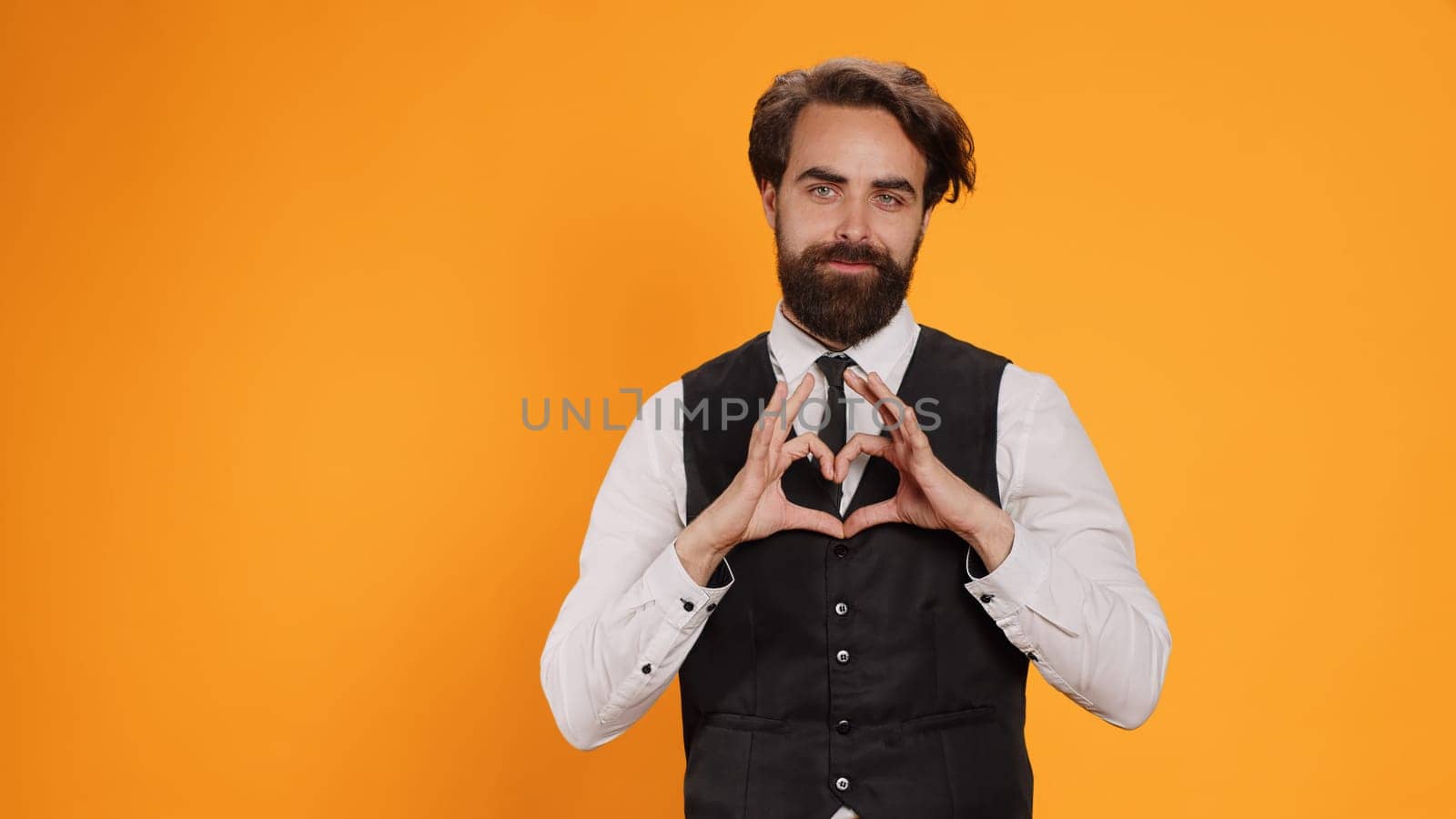 Butler indicates heart shaped sign in studio, working at five star restaurant and expressing honest emotions. Friendly employee acting romantic and presenting feelings with sweet gesture.