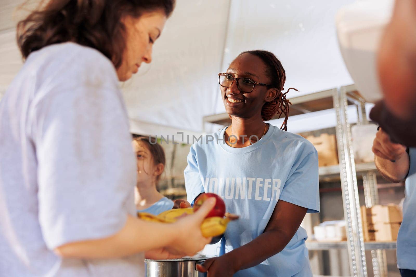 Portrait of female charity worker handing out fresh fruits and food to poor and homeless people. Cheerful black woman volunteers at food drive serving free meals to the hungry and less fortunate.