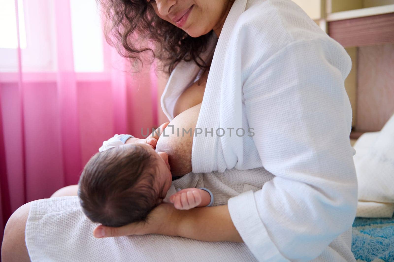 Loving caring mother in white waffle bathrobe, hugging and breastfeeding her newborn baby, sitting on the bed in cozy bedchamber interior. Lactation. Childhood and maternity lifestyle