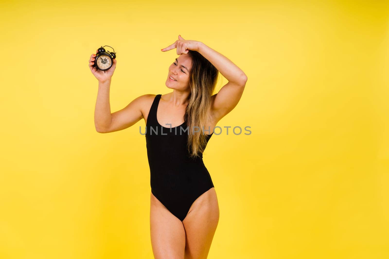 Female holding alarm clock, isolated on yellow. Young model in black swimsuit. by Zelenin