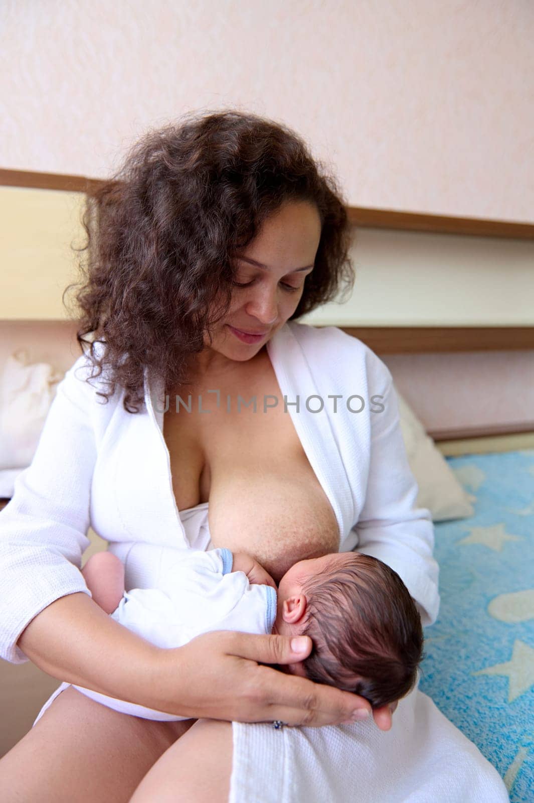 Portrait of a loving caring mother and her cute newborn baby. Happy mother sitting on the bed in cozy home interior, smiles looking at her baby boy while breastfeeding him. Tenderness. Love. Care