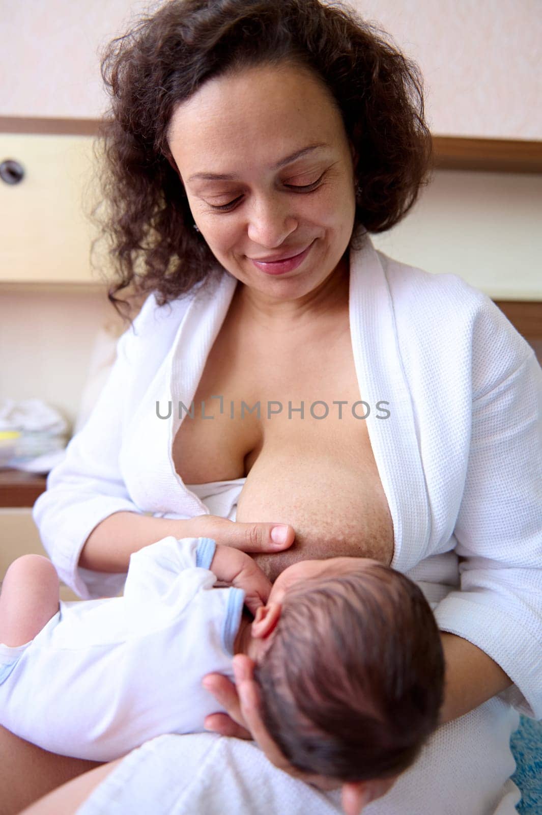 Young curly haired multi ethnic woman, loving caring mother holding and breastfeeding her newborn baby, sitting on the bed in white bedchamber interior. Family relationships. Maternity lifestyle