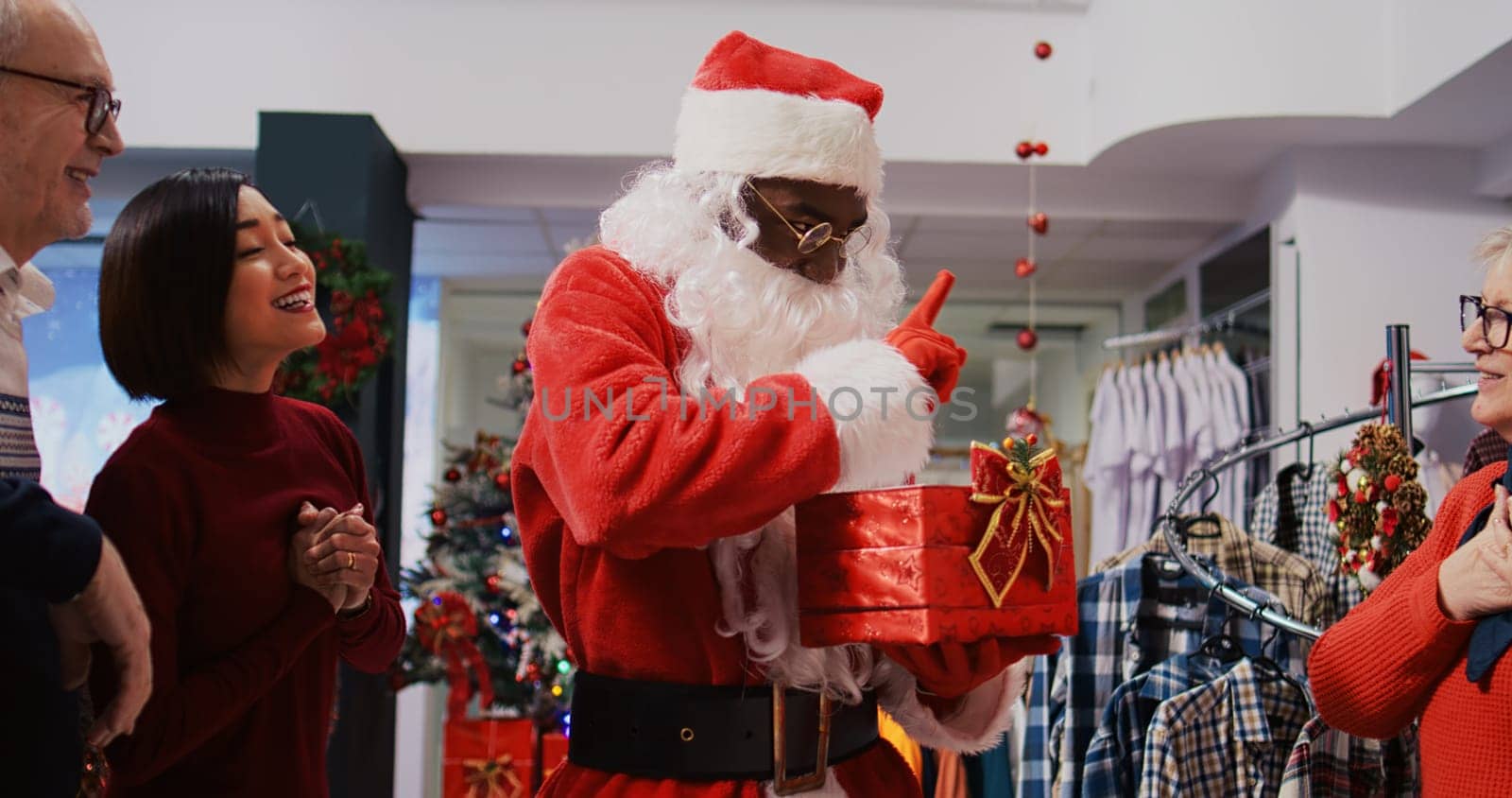 Retail assistant wearing Santa Claus suit in Christmas ornate clothing store, holding present box acting as raffle tickets container, drawing random asian woman as winner among excited clients