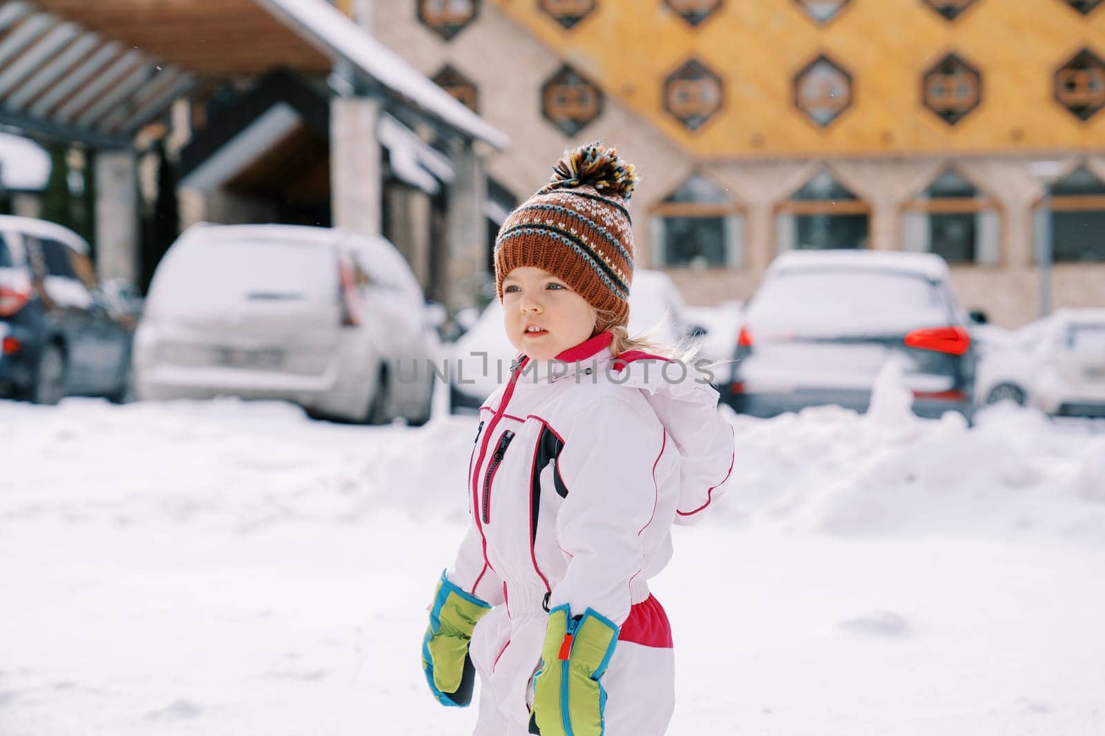 Little girl stands sideways in the snow near cars parked near the hotel. High quality photo