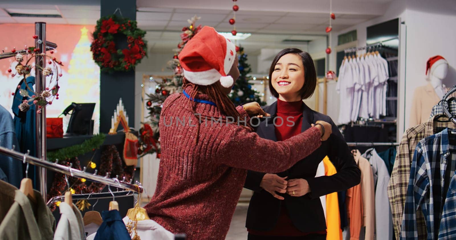 Asian client getting out of changing room, excited about elegant blazer fitting perfectly, speaking with helpful retail assistant in Christmas themed clothing store during winter holiday season