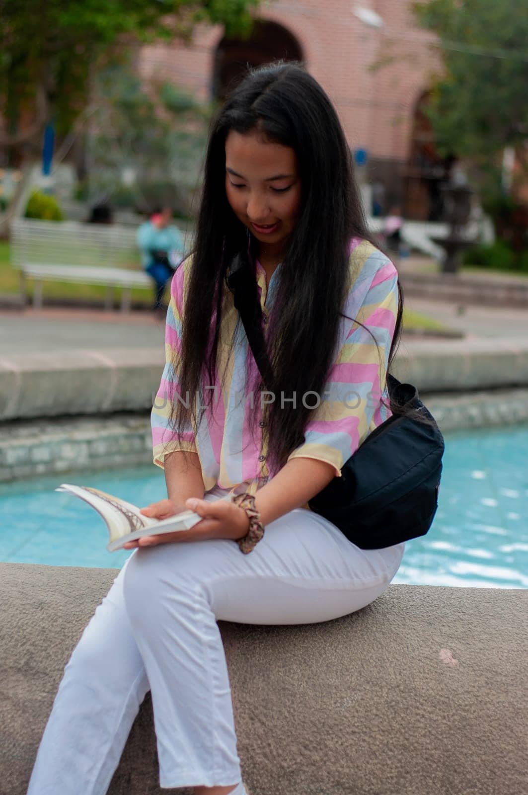 latina teenager with long black hair sitting reading a book at a fountain in a square. book day. High quality photo