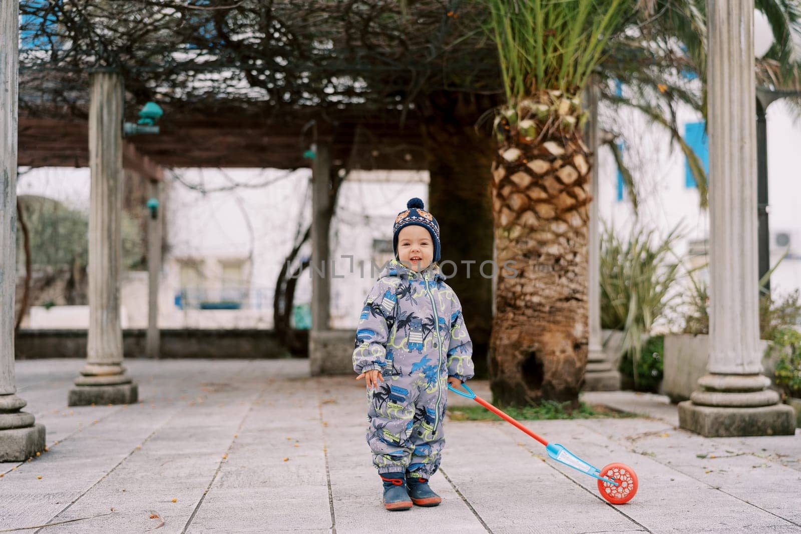 Little smiling girl with a ratchet on a stick stands near a pergola column. High quality photo