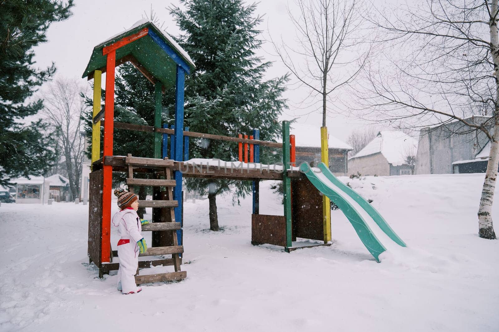 Little girl stands near a snow-covered colorful slide among the trees and looks to the side by Nadtochiy