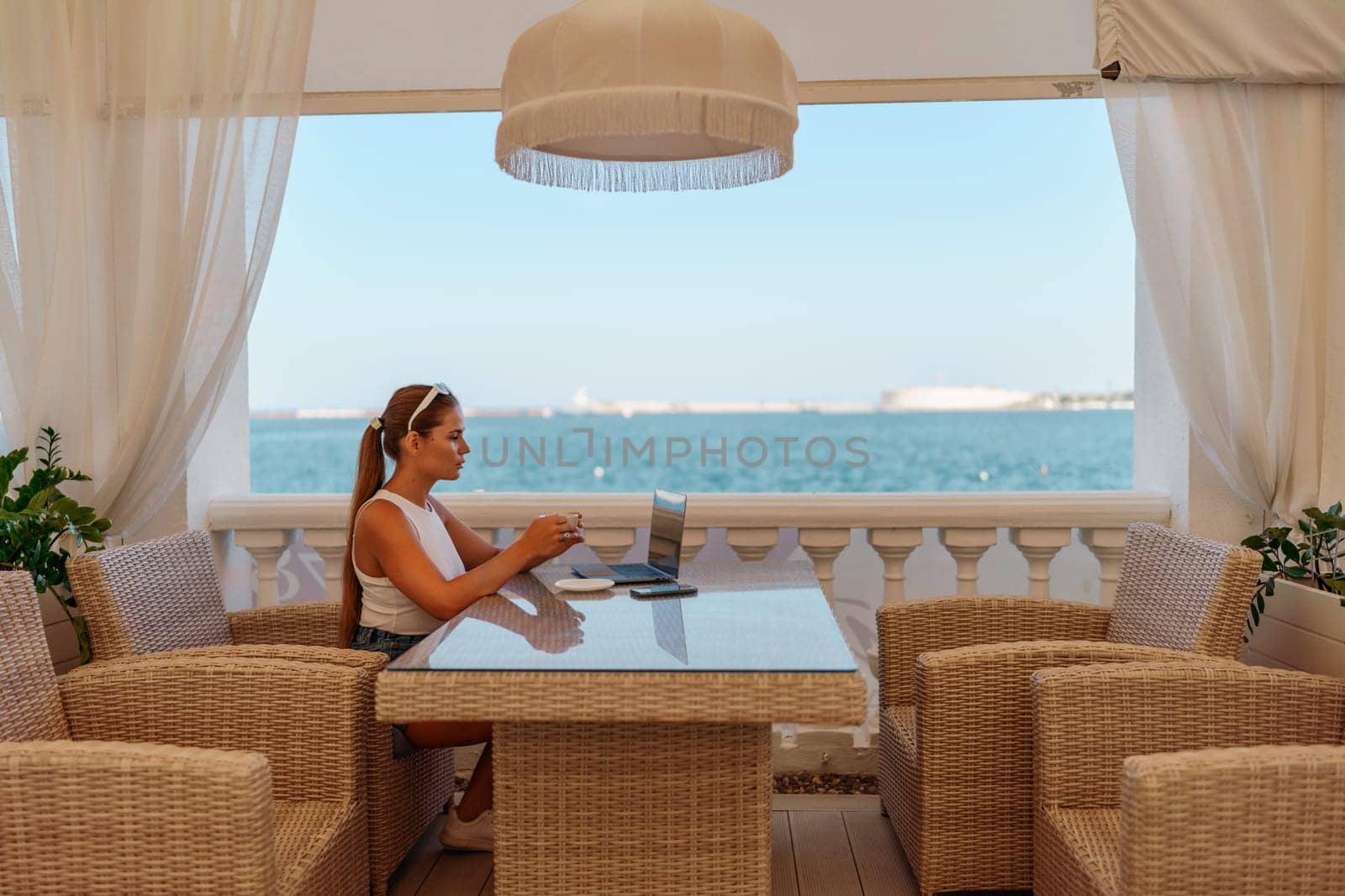 Woman coffee cafe laptop sea. Female freelancer using laptop on coffee break in modern cafe with sea view in the morning.