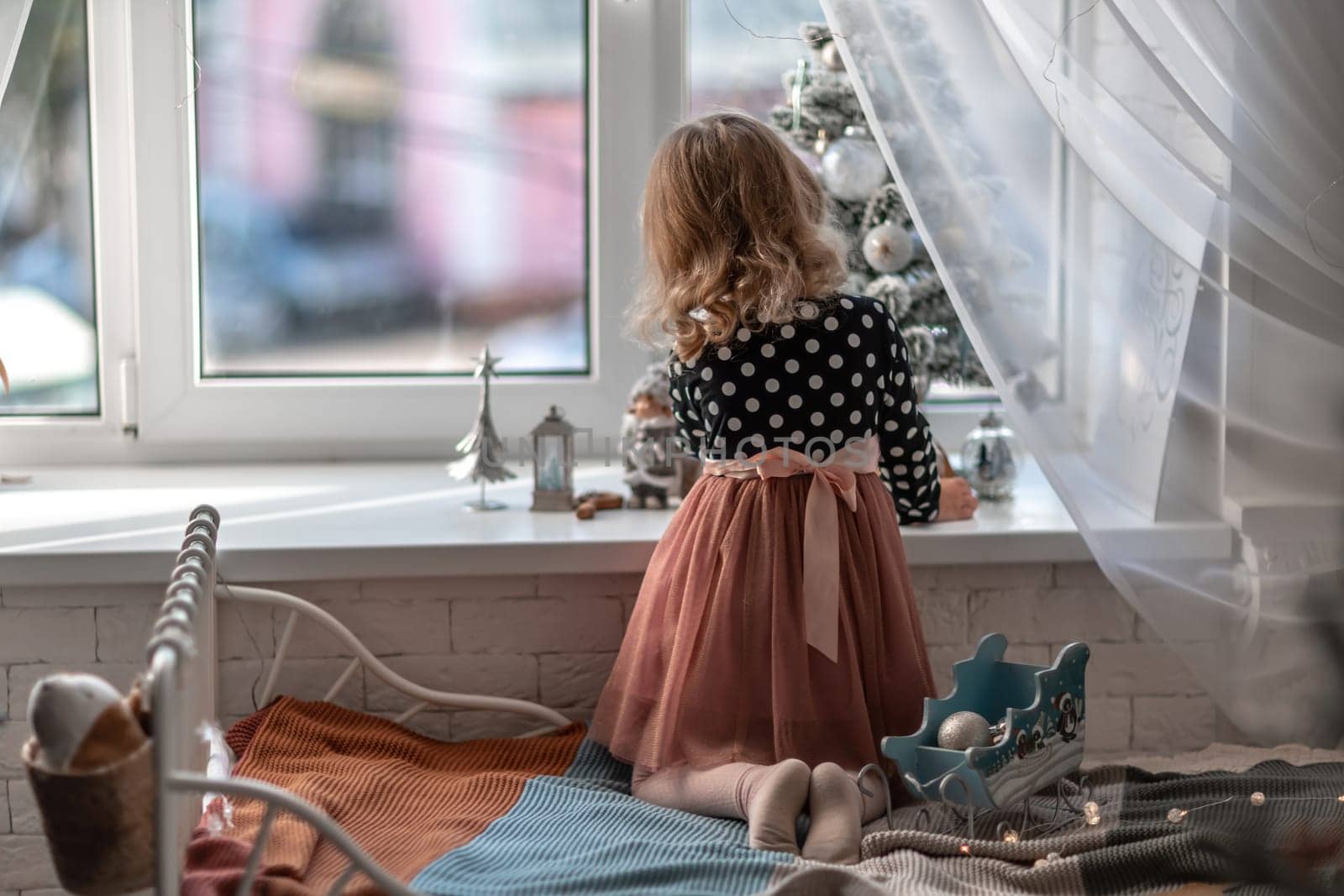 A little girl is sitting on the bed by the window and decorating a small tree with tiny Christmas toys. Happy healthy child celebrating a traditional family holiday. Adorable baby