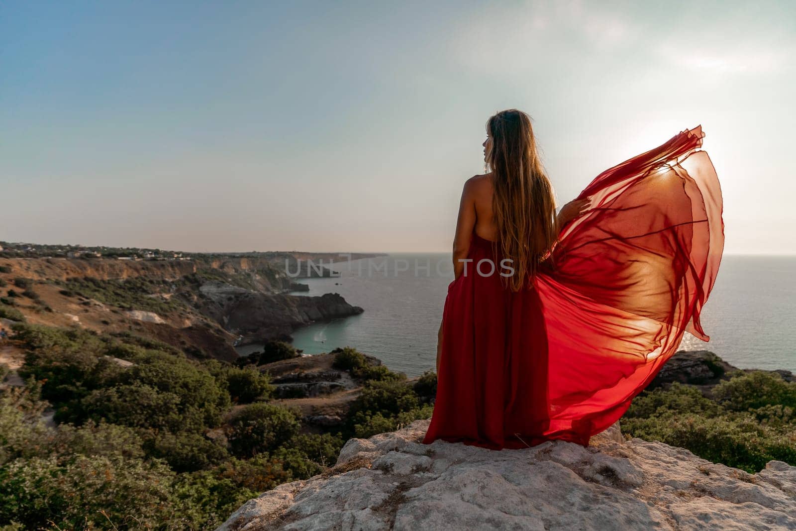 Woman sunset sea red dress, back view a happy beautiful sensual woman in a red long dress posing on a rock high above the sea on sunset. by Matiunina