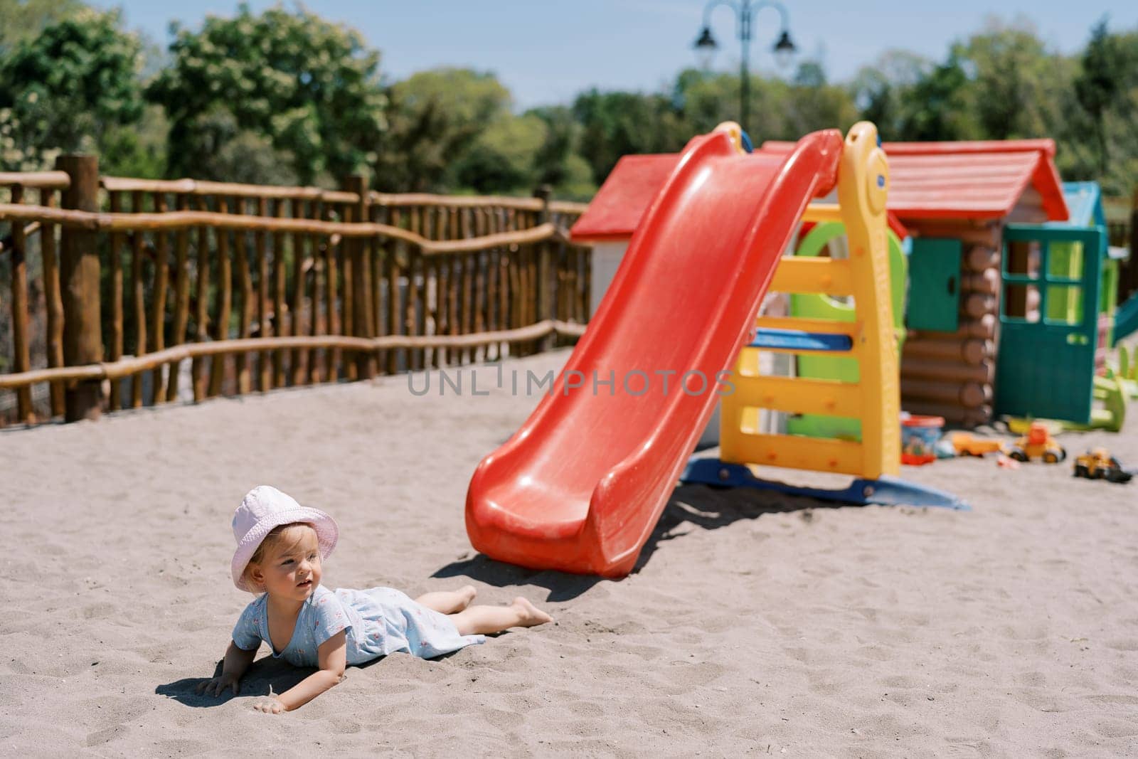Little girl lies on the sand near a slide on the playground and looks away. High quality photo