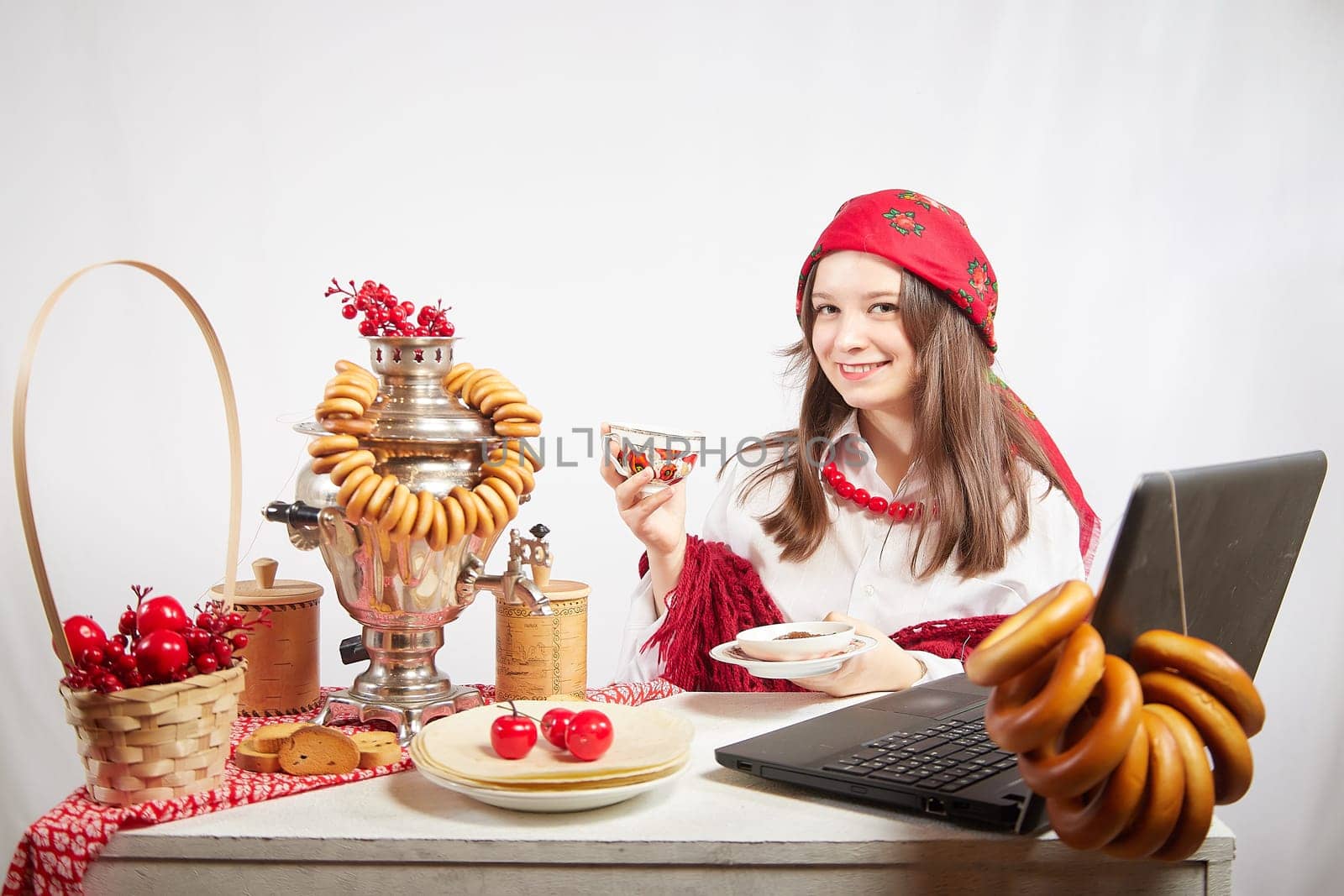 Girl in red colored scarf, white shirt and shawl near a table with a samovar, bagels drinks tea for the Easter holiday and a Masolennitsa. A freelancer or blogger with a laptop is resting and working by keleny