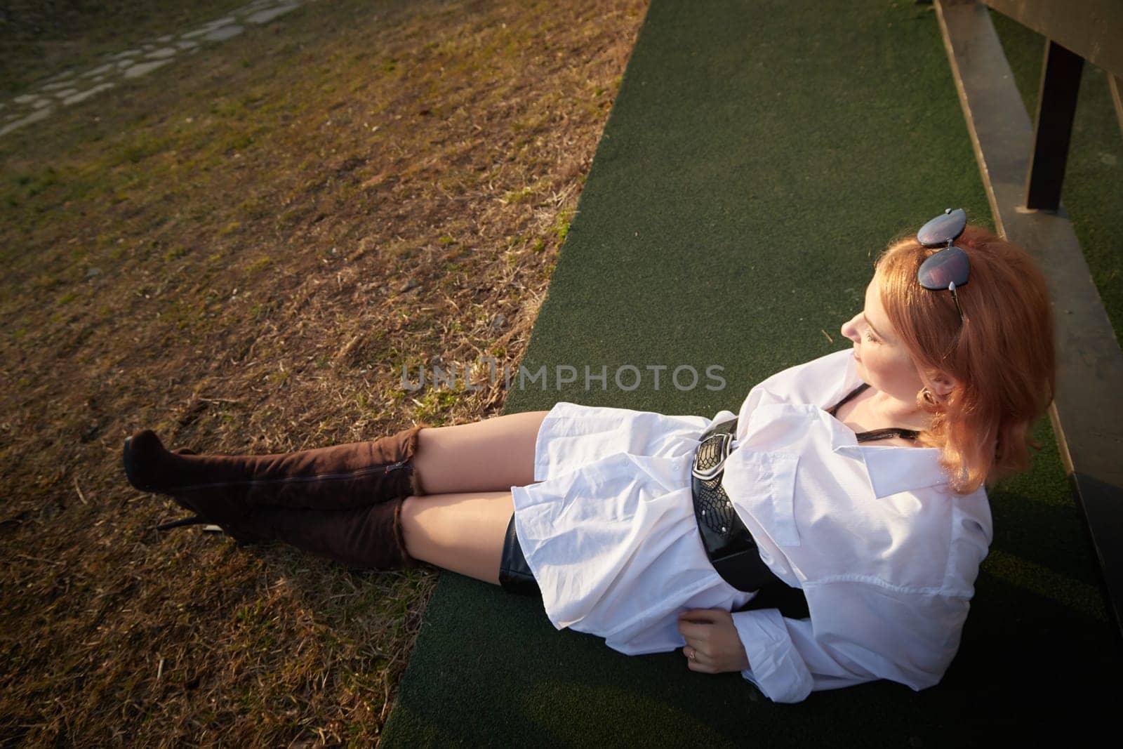 Beautiful girl with red hair in white shirt in open wooden pavillion in village or small town. Young slender woman and sky with clouds on background on autumn, spring or summer day