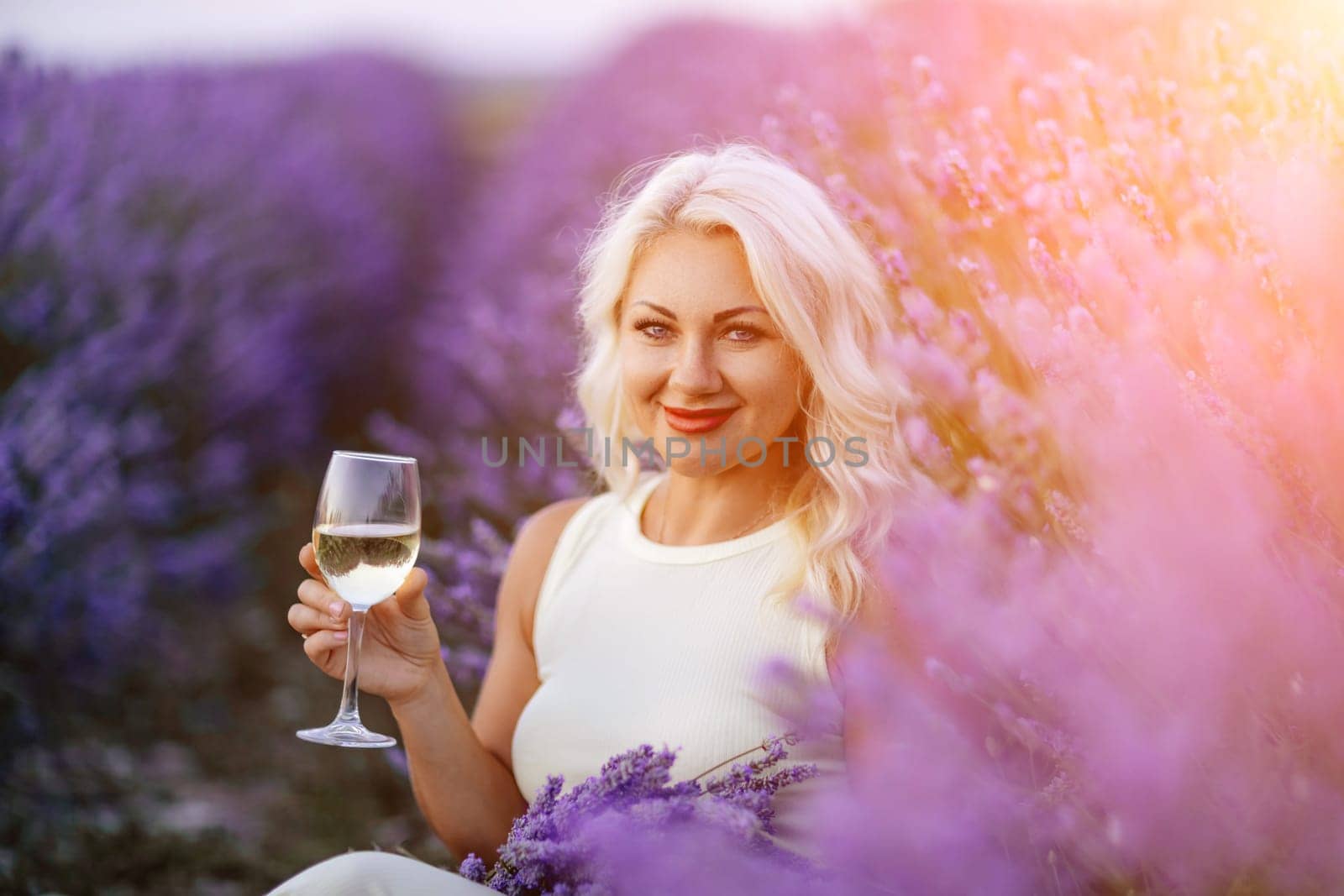 Blonde lavender field holds a glass of white wine in her hands. Happy woman in white dress enjoys lavender field picnic holding a large bouquet of lavender in her hands . Illustrating woman's picnic in a lavender field. by Matiunina