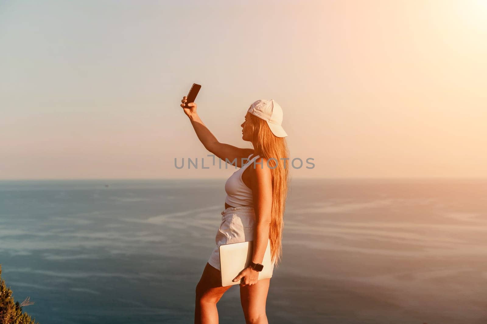 Selfie woman sea. The picture depicts a woman in a cap and tank top, taking a selfie shot with her mobile phone, showcasing her happy and carefree vacation mood against the beautiful sea backgroun.