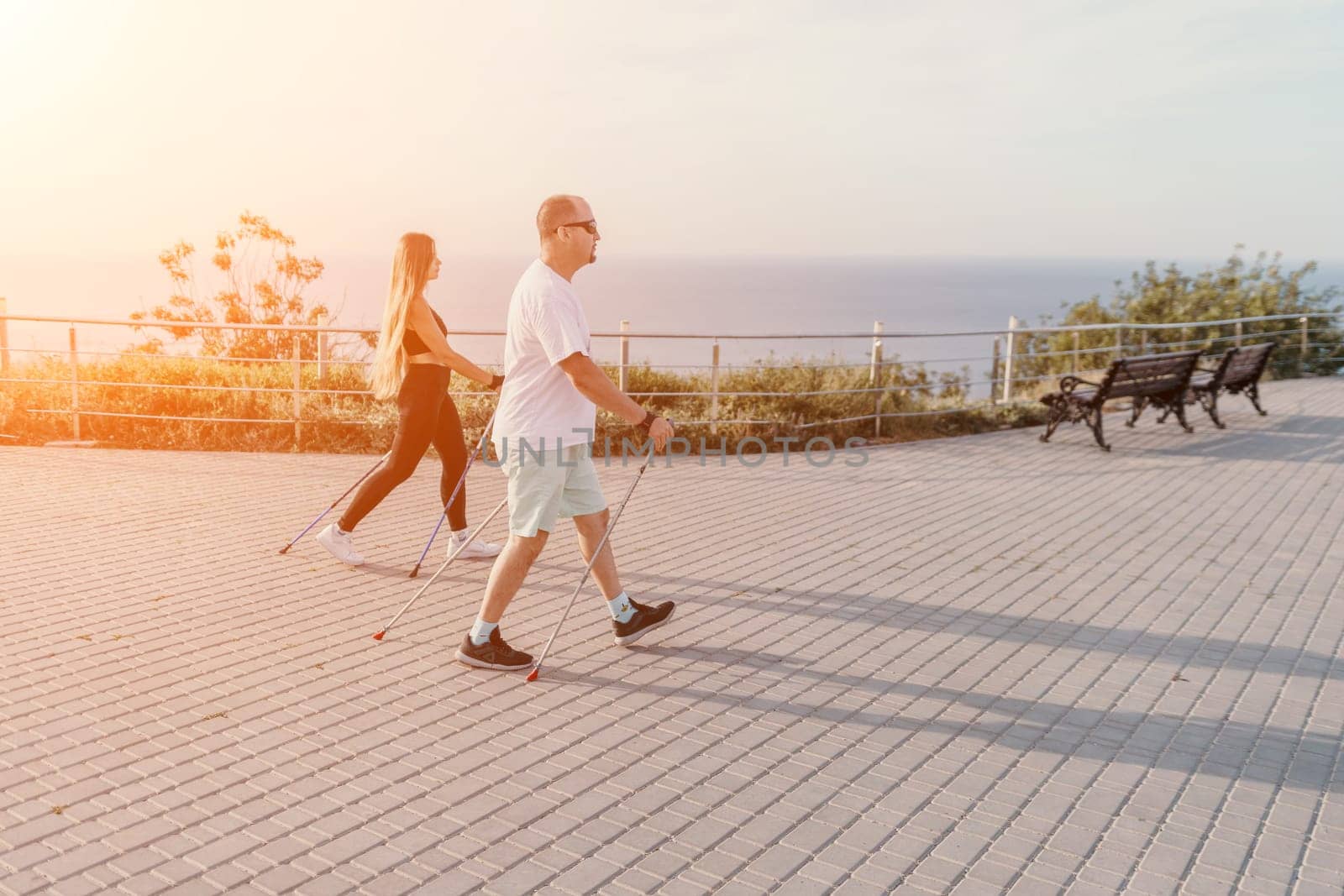 Happy Middle aged couple or friends practicing nordic walking in park near sea. Mature couple with trekking poles walking, practicing Nordic walking outdoors. Aging youthfully and sport concept.