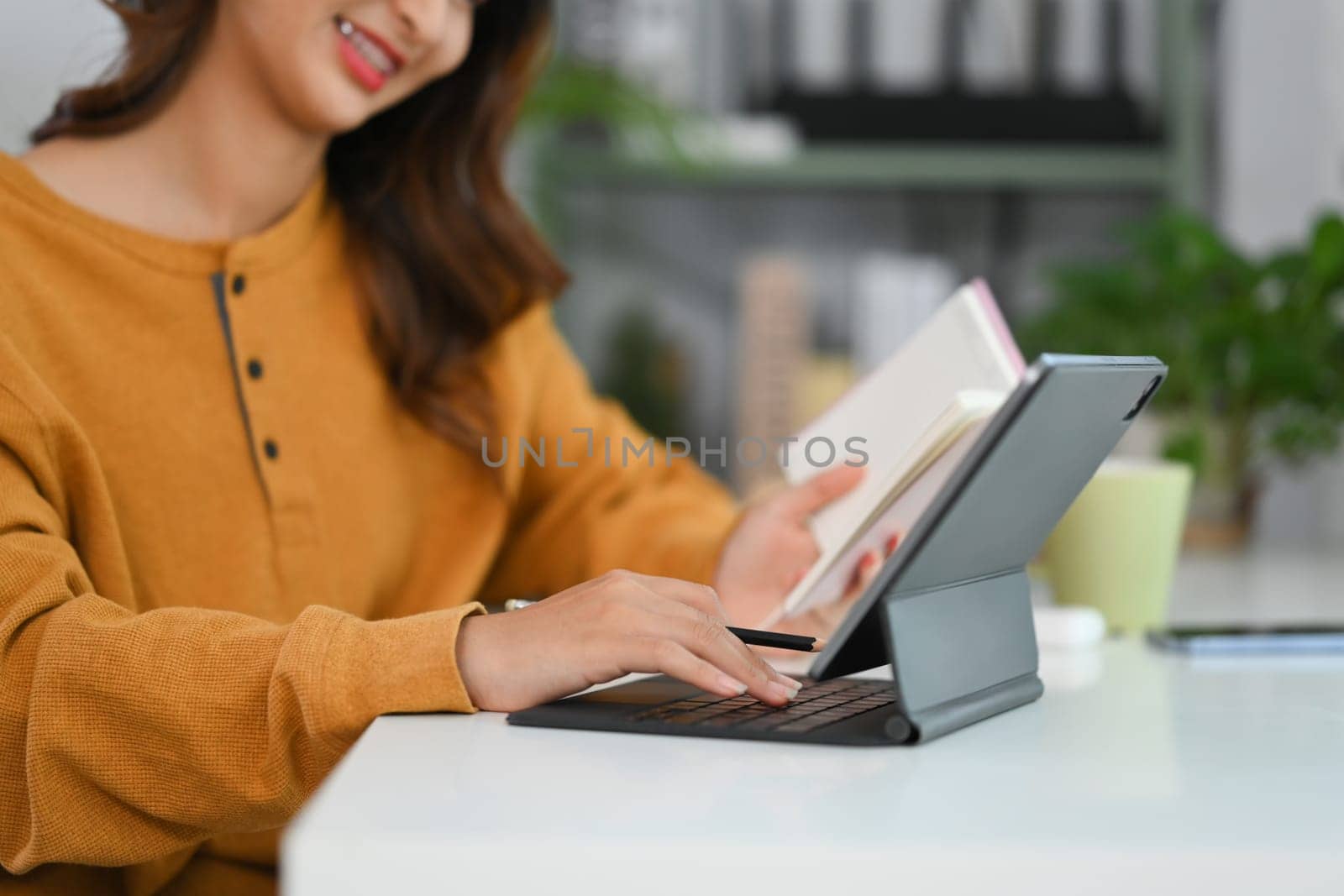 Happy young woman holding notebook and typing in keyboard of digital tablet