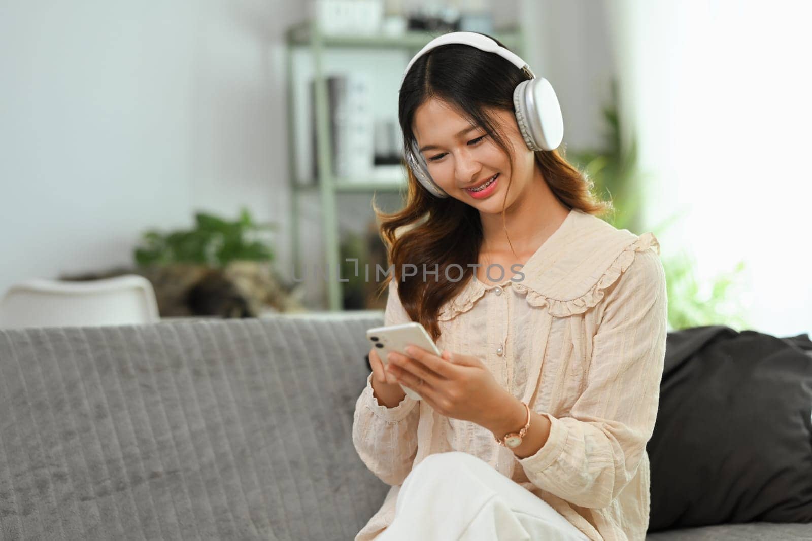 Cheerful young woman wearing headphone using mobile phone on couch at home. by prathanchorruangsak