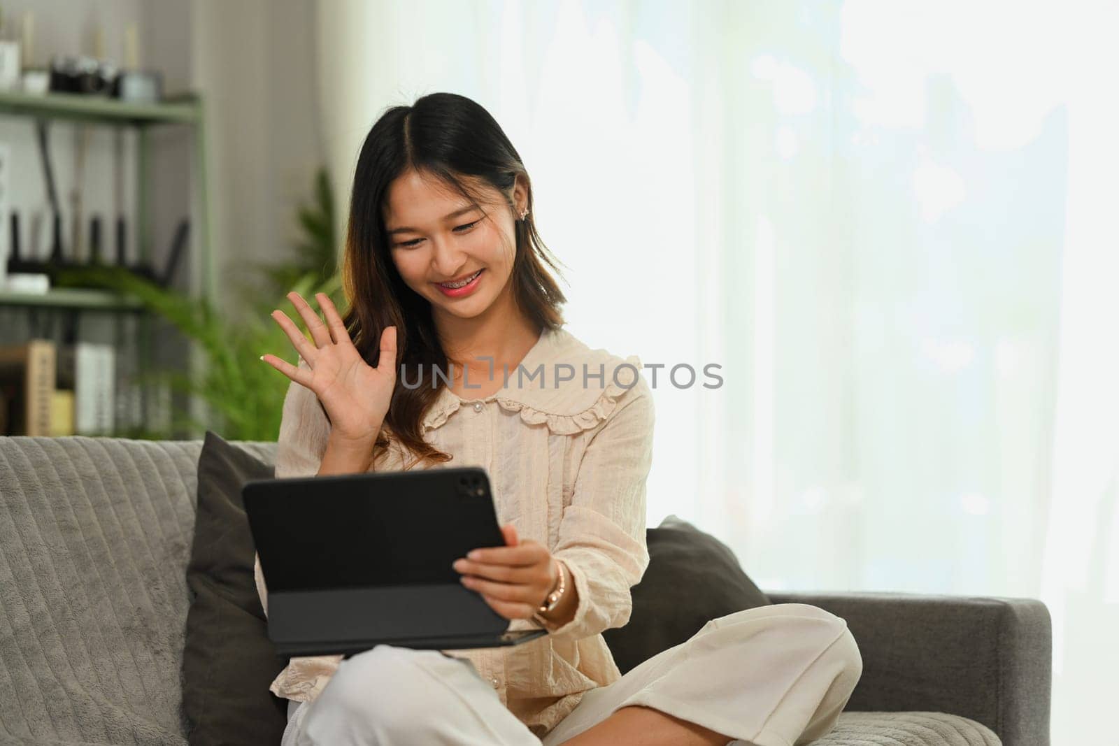 Smiling young woman in casual clothes waving hand while making video call via digital tablet. by prathanchorruangsak