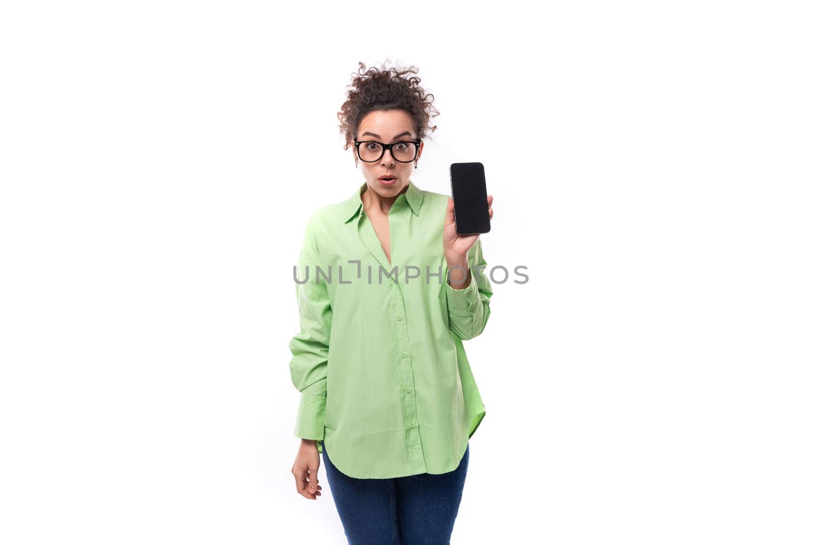 young pretty european brunette curly woman dressed in a light green shirt and glasses holds a smartphone.