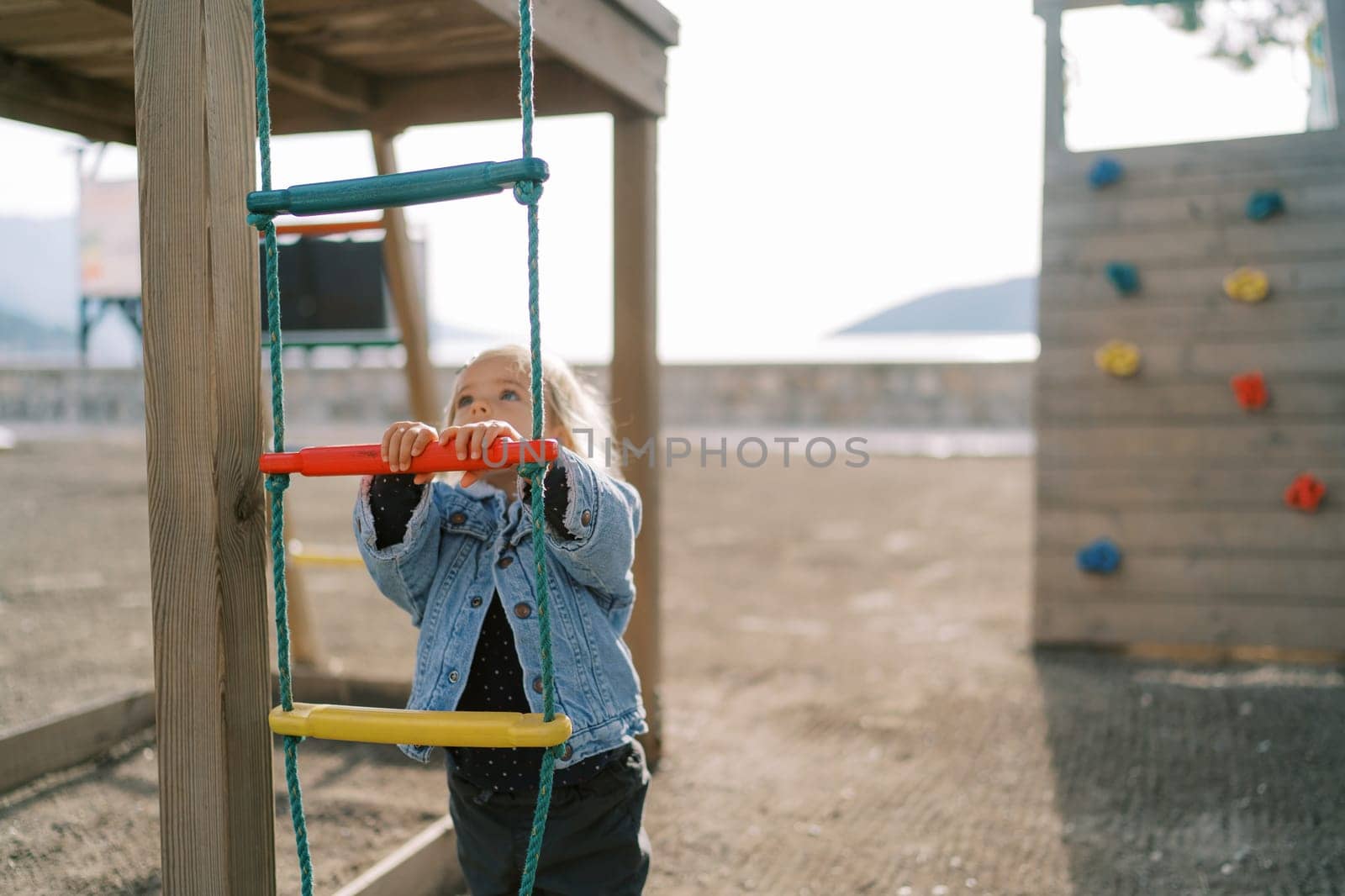 Little girl stands near a rope ladder on a playground and looks up by Nadtochiy