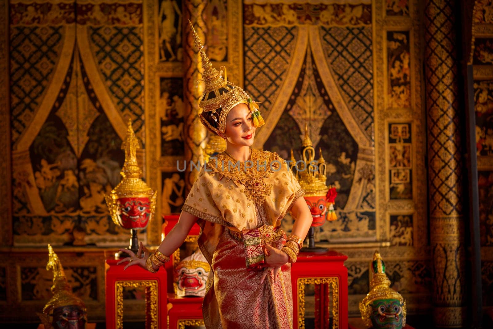 Beautiful Asian woman wear Thai ancient traditional dress action of dancing in front of Khon or traditional Thai classic masked from the Ramakien on wooden shelves and painting on public place wall.