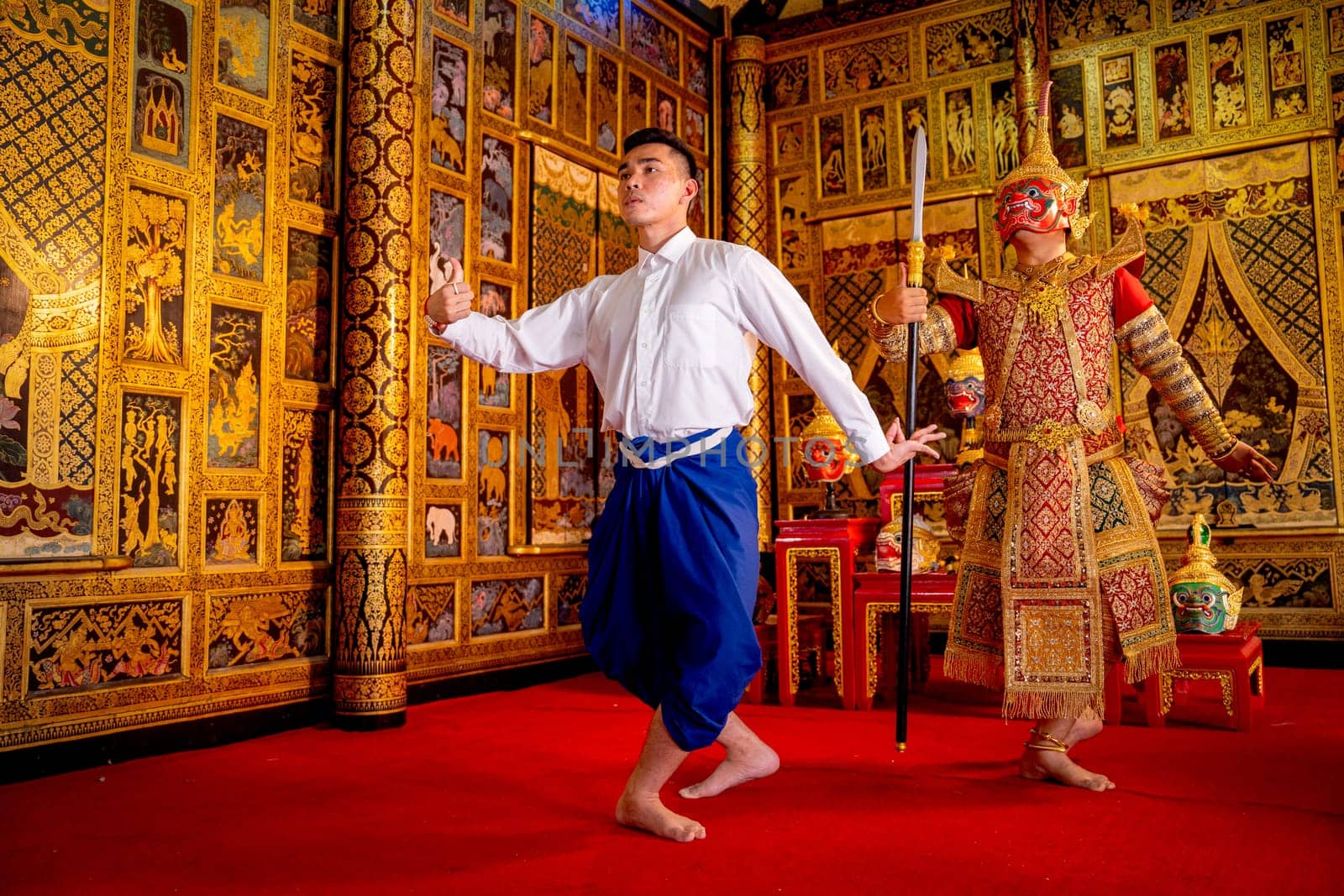 Asian man with Thai style cloth stand in front of man wear ancient traditional Thai pattern Pantomime also action to teach the other man in public place with Thai painting style on wall.