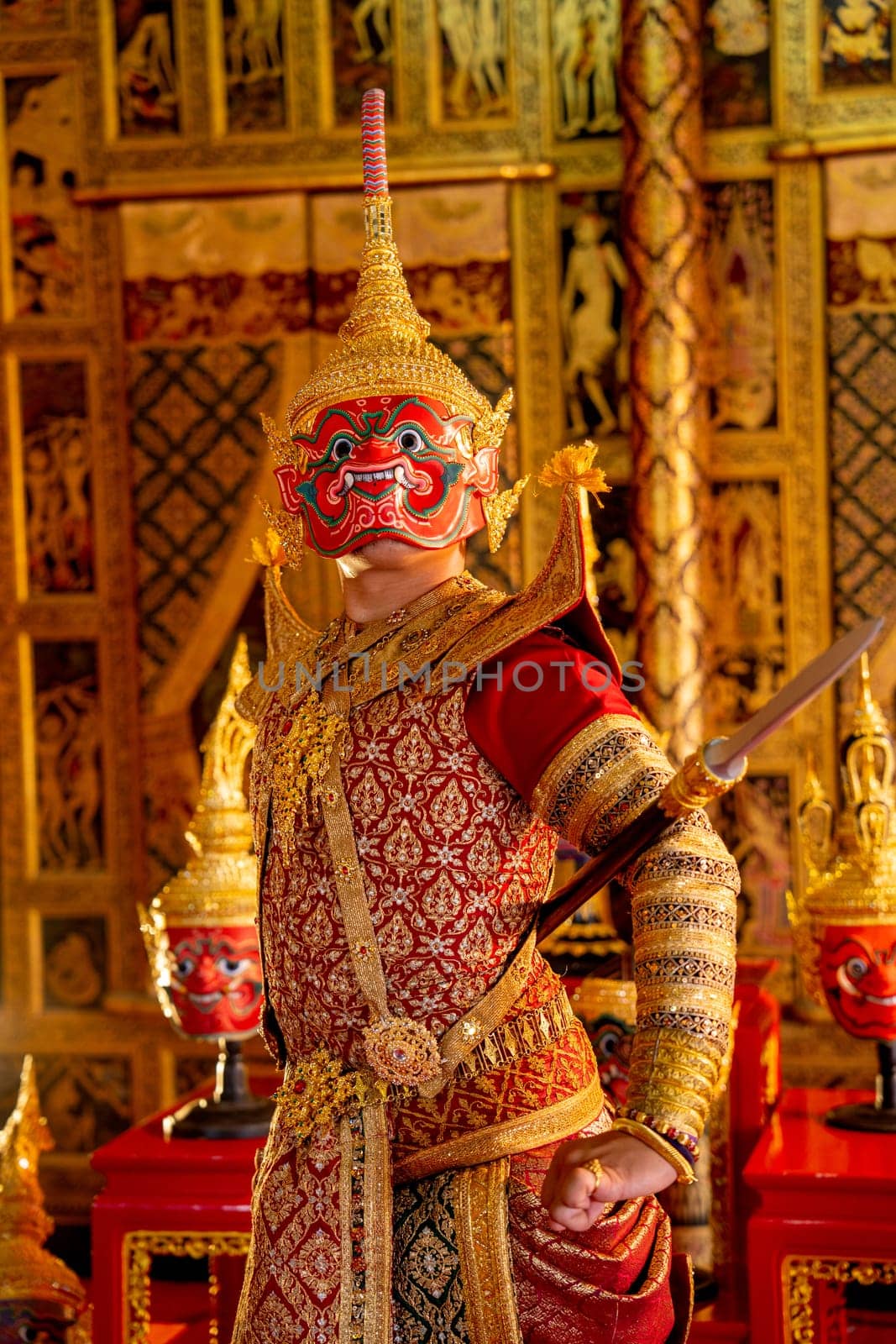 Close up Khon or traditional Thai classic masked from the Ramakien with Red giant character stand with spear and look forward with Thai painting wall in background.