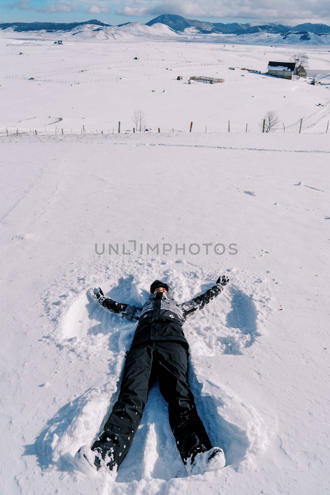 Young woman in a ski suit makes a snow angel in the snow by Nadtochiy