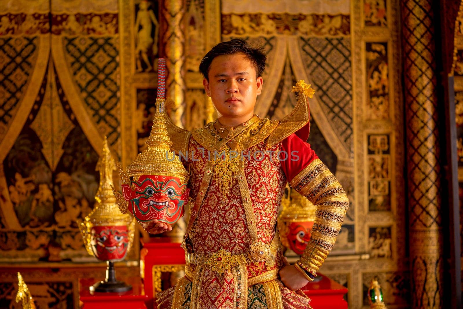 Portrait of Asian man hold Pantomime or Khon masks with red giant character and look at camera also stay in front of Thai painting on wall in public place.