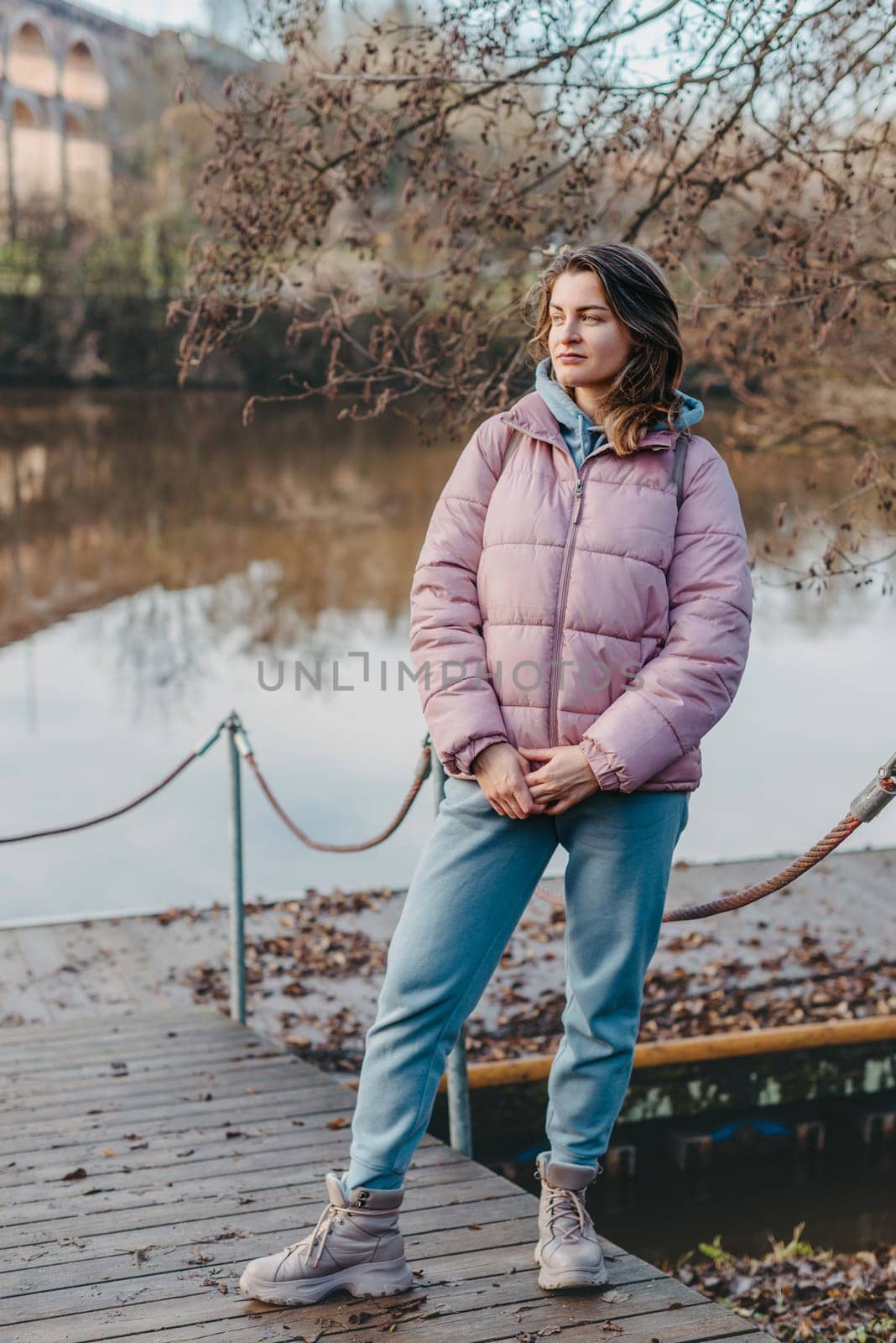 A young woman standing at the shore looking at the river in autumn sunny day. Street view, copy space for text, travel photo. Happy tourist woman on the bank of the river in autumn in warm clothes. Tourists enjoy their vacation, winter season. Romantic look and travel concept. A joyful mood in a caucasian girl. Winter Wonderland: Enchanting Girl by the Riverside in Autumn by Andrii_Ko