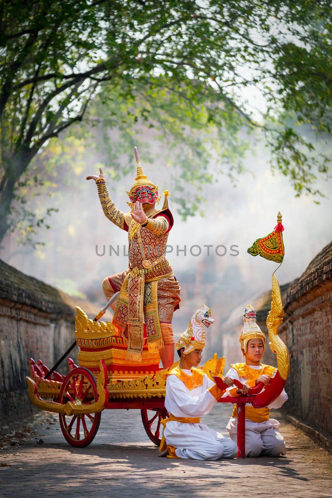 Vertical image of Khon or traditional Thai classic masked from the Ramakien as character of red giant stand and dance on traditional chariot also hold weapon stay in front of ancient building.