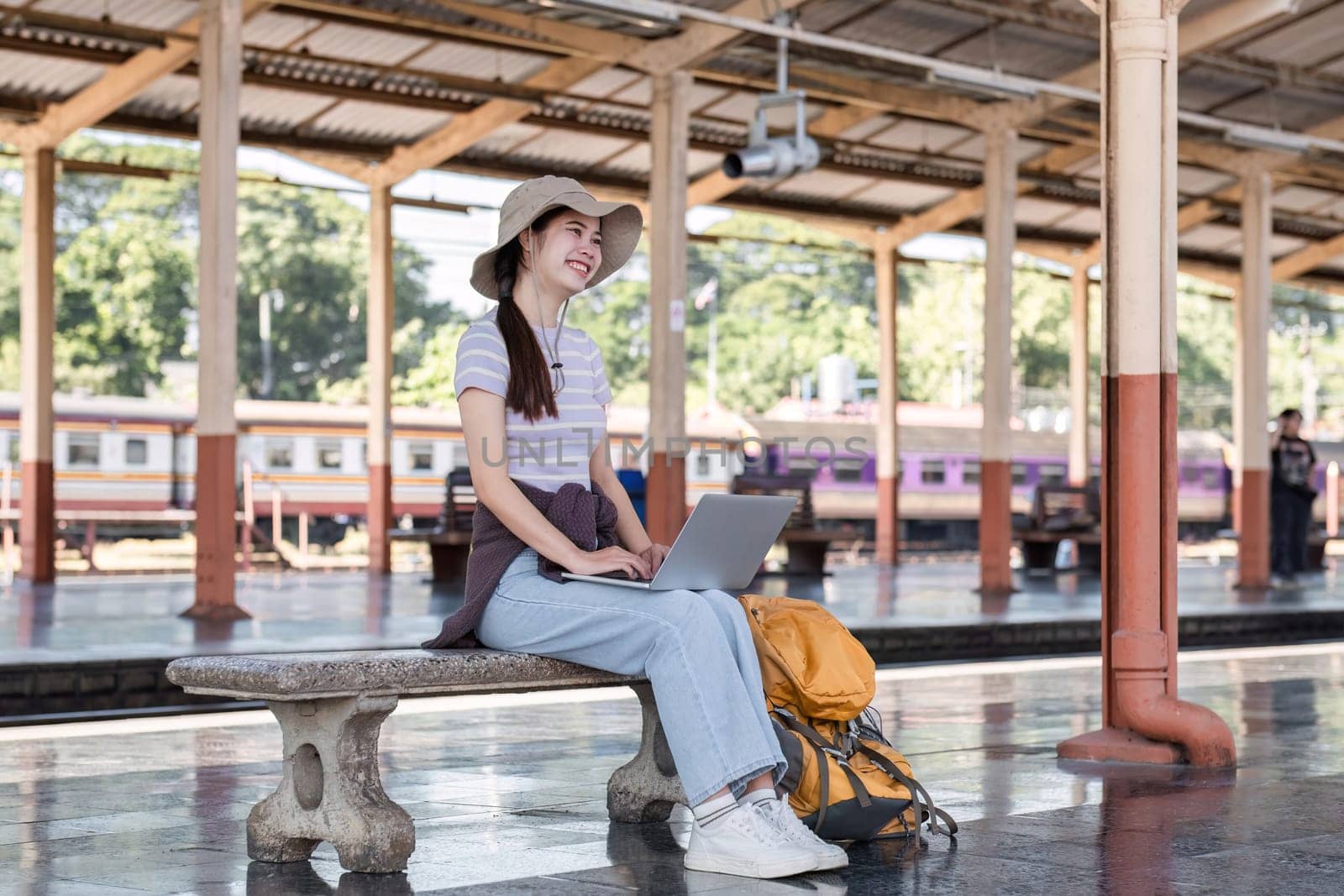 Young Asian woman in modern train station Female backpacker passenger sitting on a bench using a laptop while waiting for a train at the train station to go on holiday. by wichayada