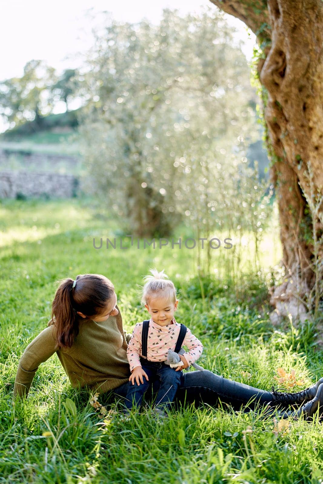 Mom looks at a little girl sitting on her lap near a tree by Nadtochiy