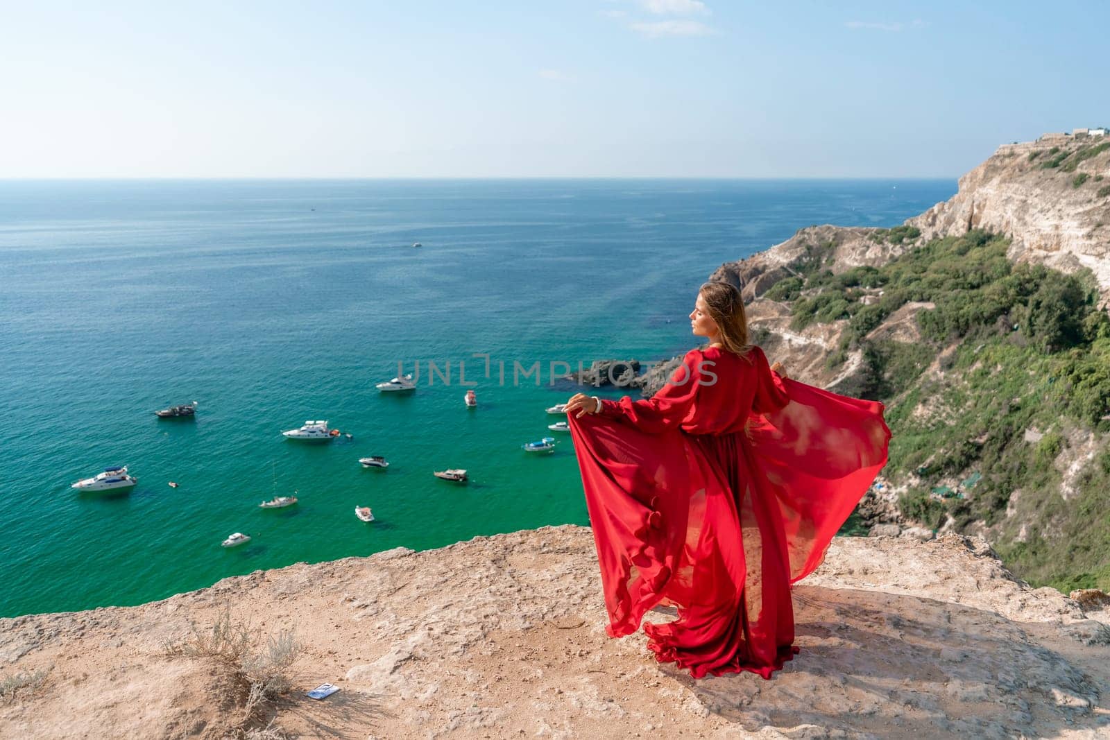 Red Dress Woman sea Cliff. A beautiful woman in a red dress and white swimsuit poses on a cliff overlooking the sea on a sunny day. Boats and yachts dot the background