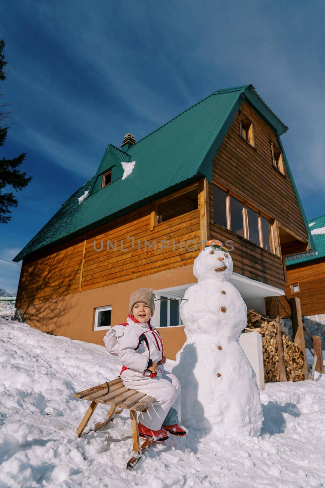 Little girl sits sideways, opening her mouth on a sleigh near a snowman in the yard by Nadtochiy