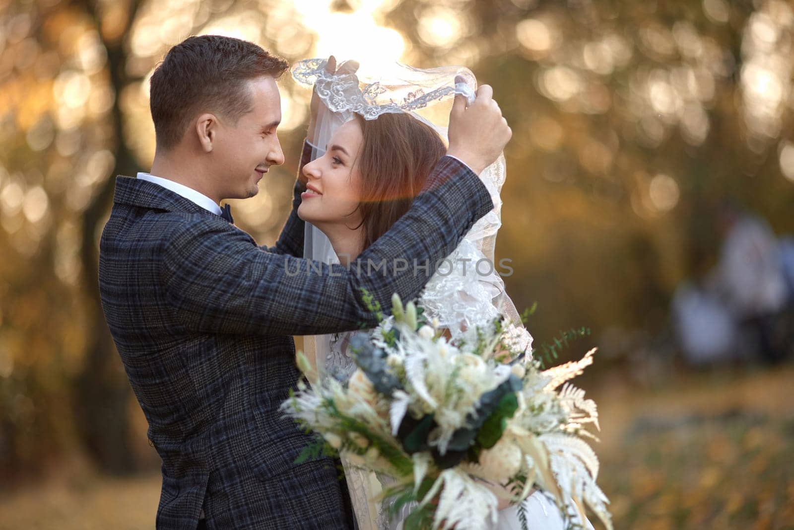 happy bride and groom enjoying romantic moments under veil and looking at each other on natural background in sunny day