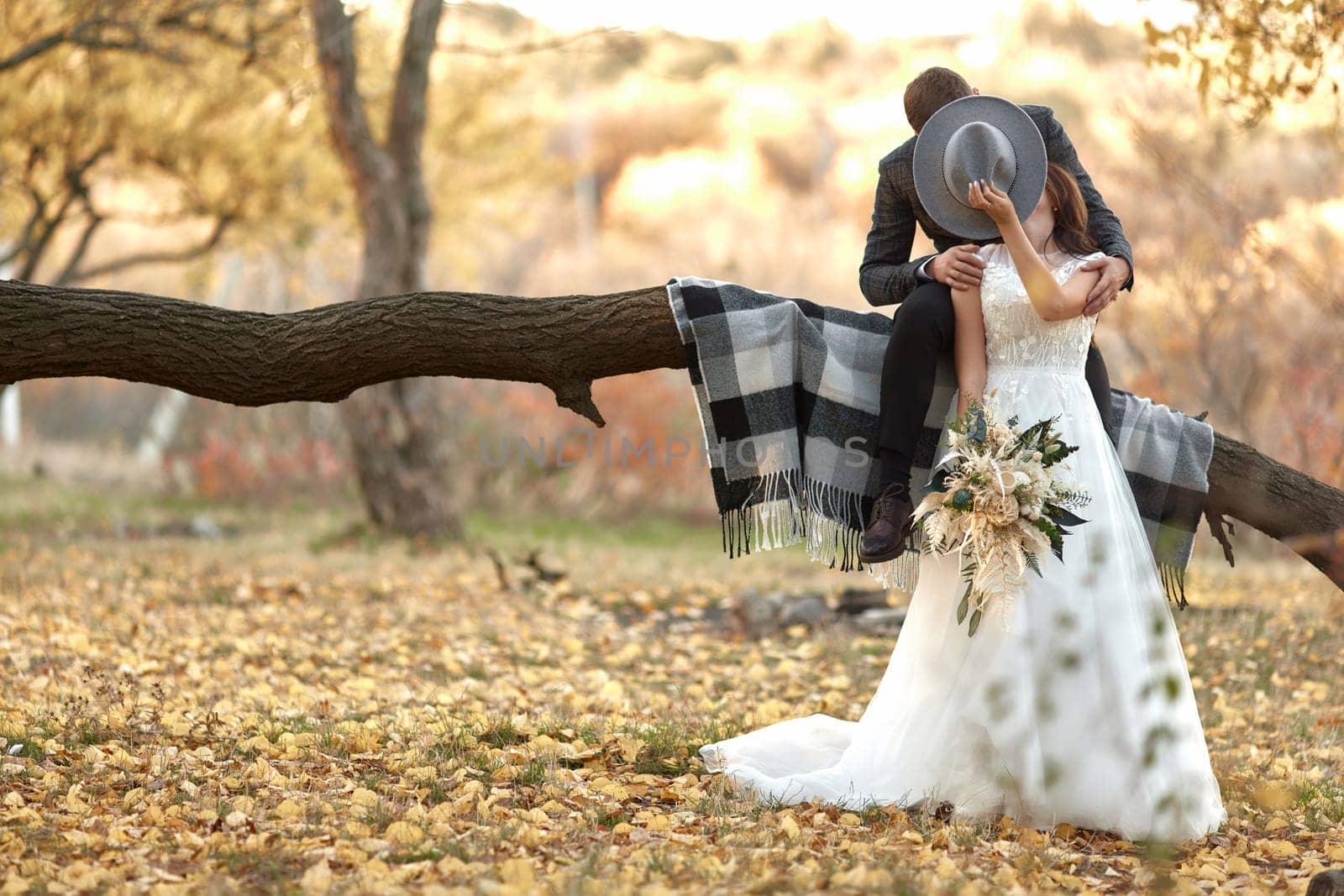 beautiful happy stylish bride and groom kissing and covering by hat near tree outdoor in autumn