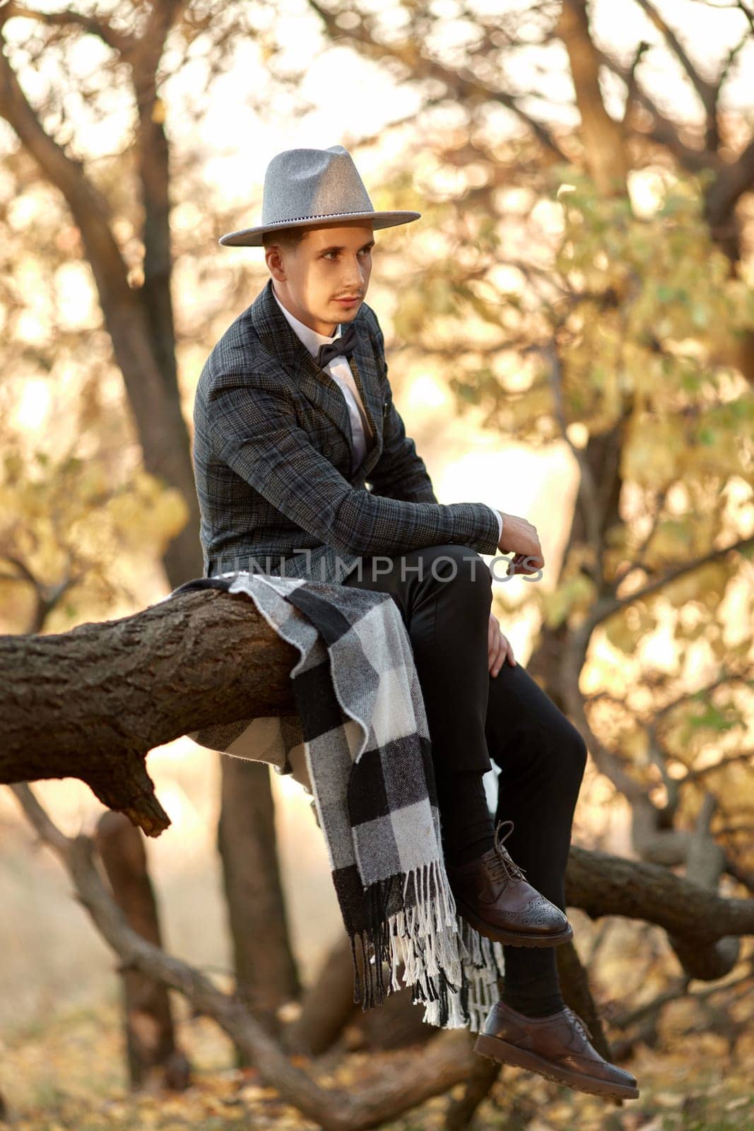 stylish guy in hat sitting on tree outdoor in autumn