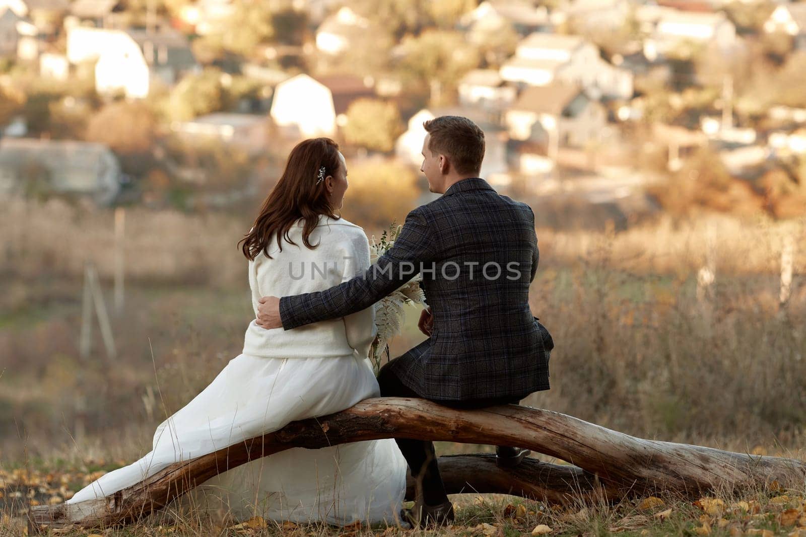 bride in white wedding dress and groom are sitting on tree in nature, back view