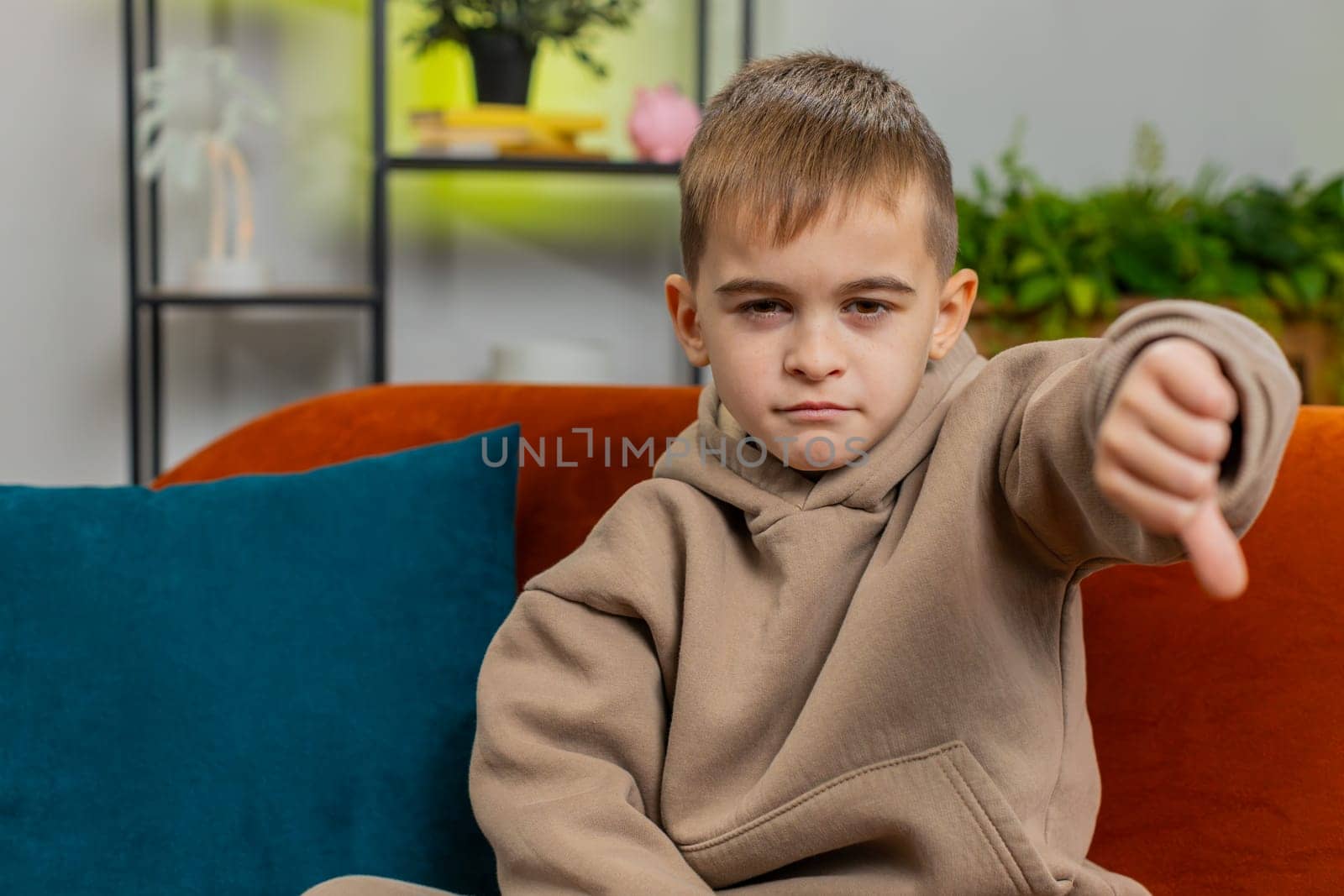 Dislike. Upset child boy showing thumbs down sign gesture, expressing discontent disapproval dissatisfied bad work at modern home apartment indoors. Displeased teenager kid in room on sofa. Copy-space