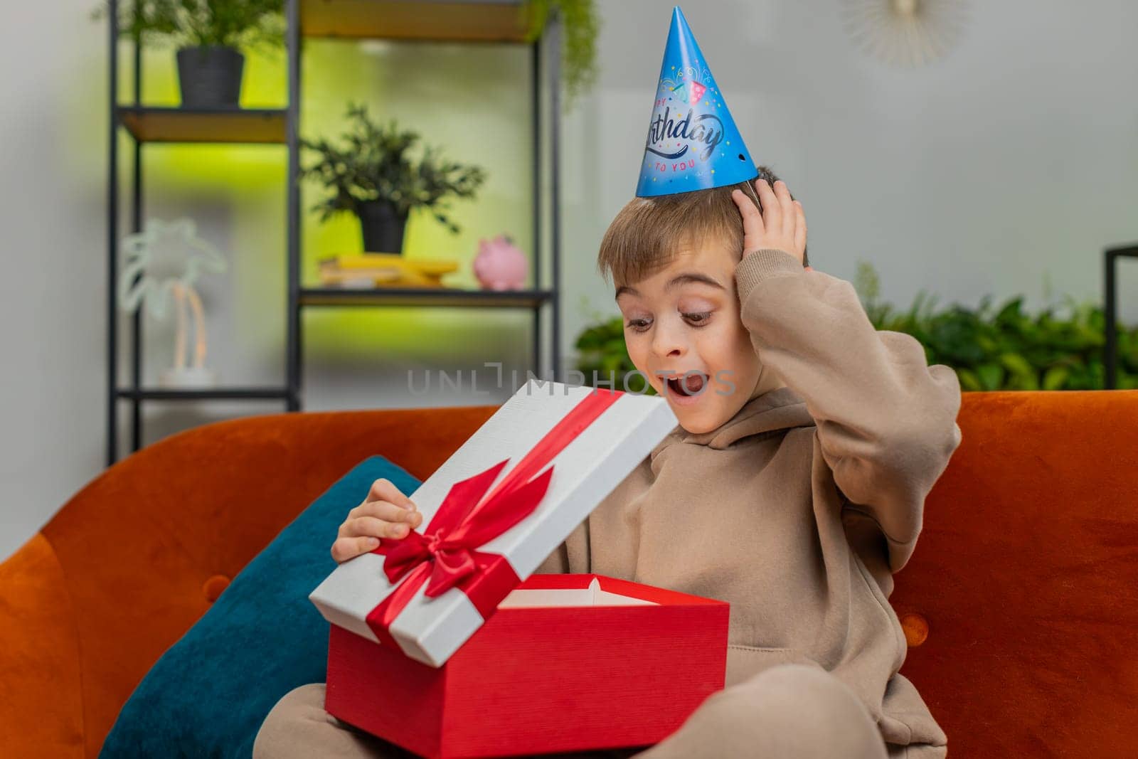 Happy preteen child boy wears festive birthday cap hat hold gift box with ribbon congratulating. Teenager male little kid celebrating party event opening delivery greeting present at home room on sofa