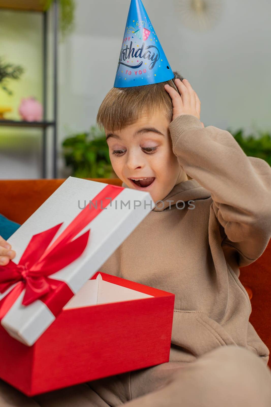 Happy preteen child boy wears festive birthday cap hat hold gift box with ribbon congratulating. Teenager kid celebrating party event opening delivery greeting present at home room on sofa. Vertical