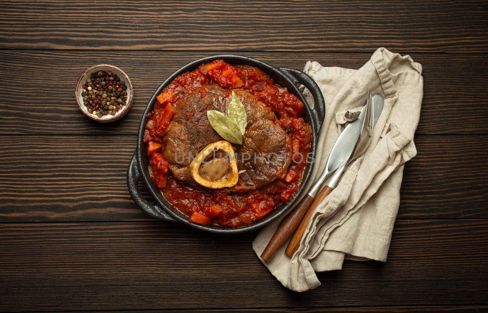 Traditional Italian dish Ossobuco all Milanese made with cut veal shank meat with vegetable tomato sauce served in black casserole pan top view on rustic brown wooden background.