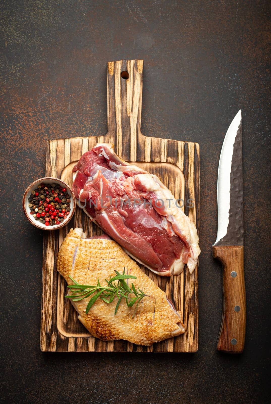 Two raw uncooked duck breast fillets with skin, seasoned with salt, pepper, rosemary top view on wooden cutting board with knife, dark brown concrete rustic background by its_al_dente