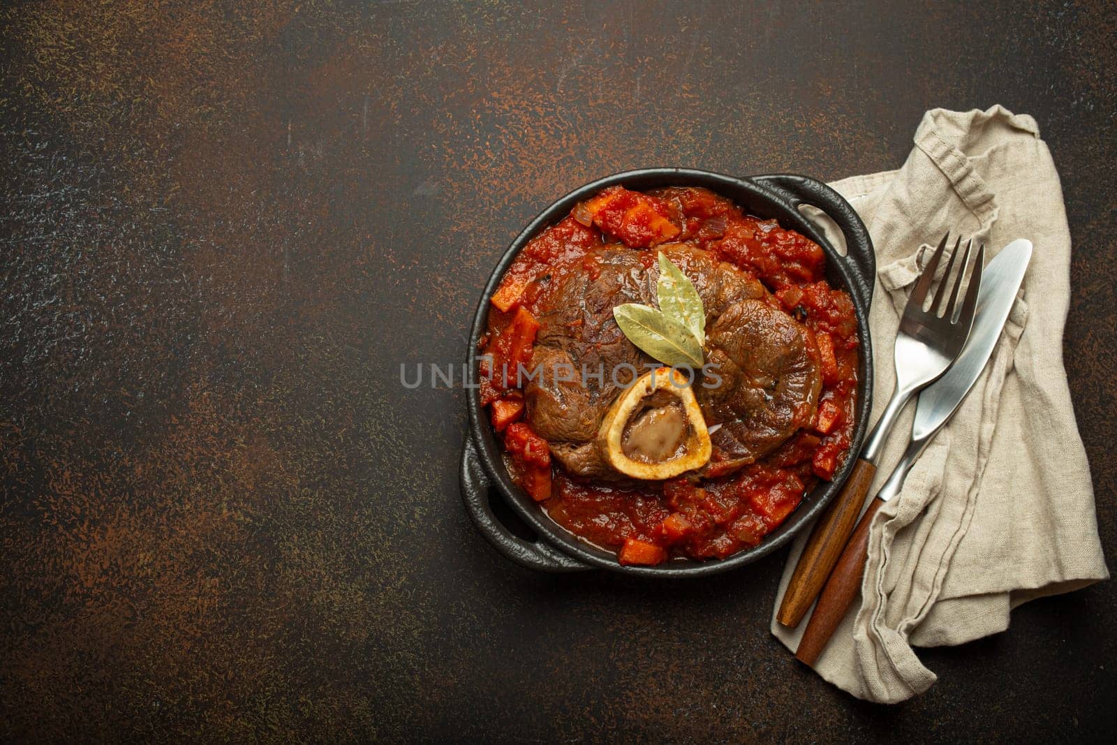 Traditional Italian dish Ossobuco all Milanese made with cut veal shank meat with vegetable tomato sauce served in black casserole pan top view on rustic brown background, copy space.